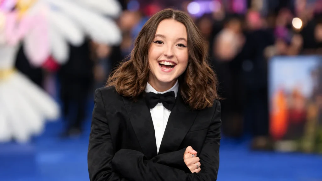 Bella Ramsey smiling in a black tuxedo with a white shirt and bow tie at a red carpet event