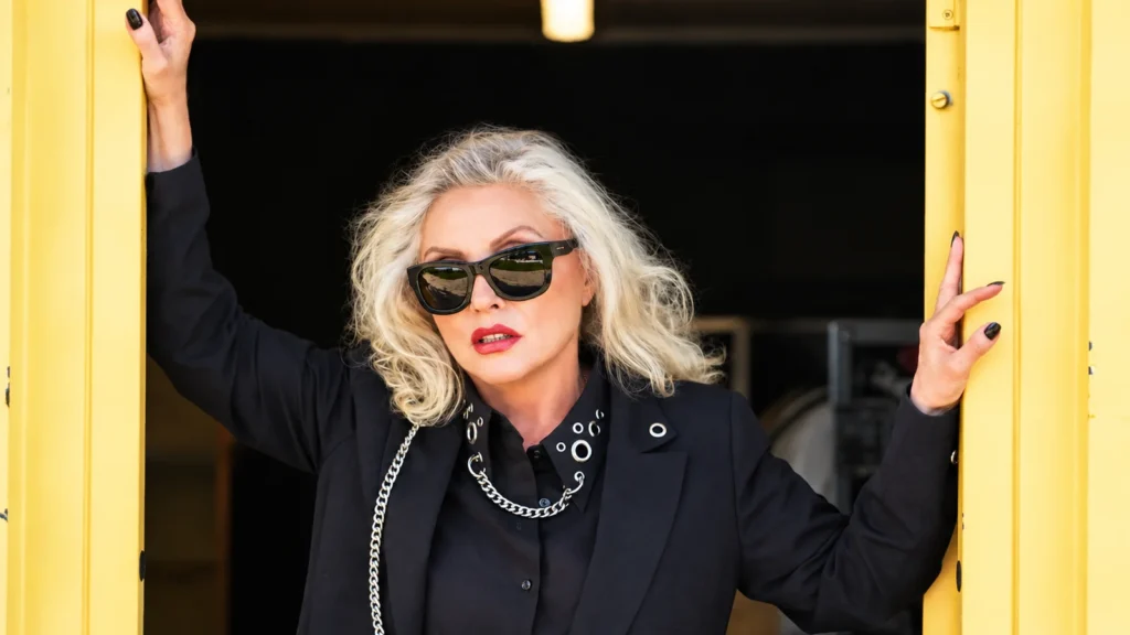 Debbie Harry wearing sunglasses and a black outfit in a yellow doorway