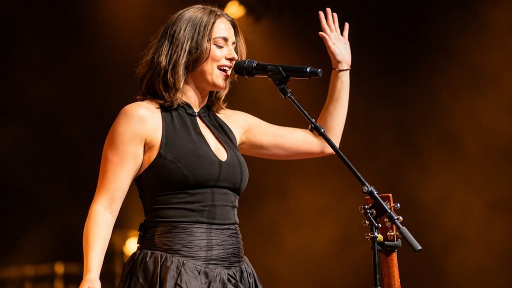 Allie Sherlock singing live on stage in a black dress under warm concert lighting