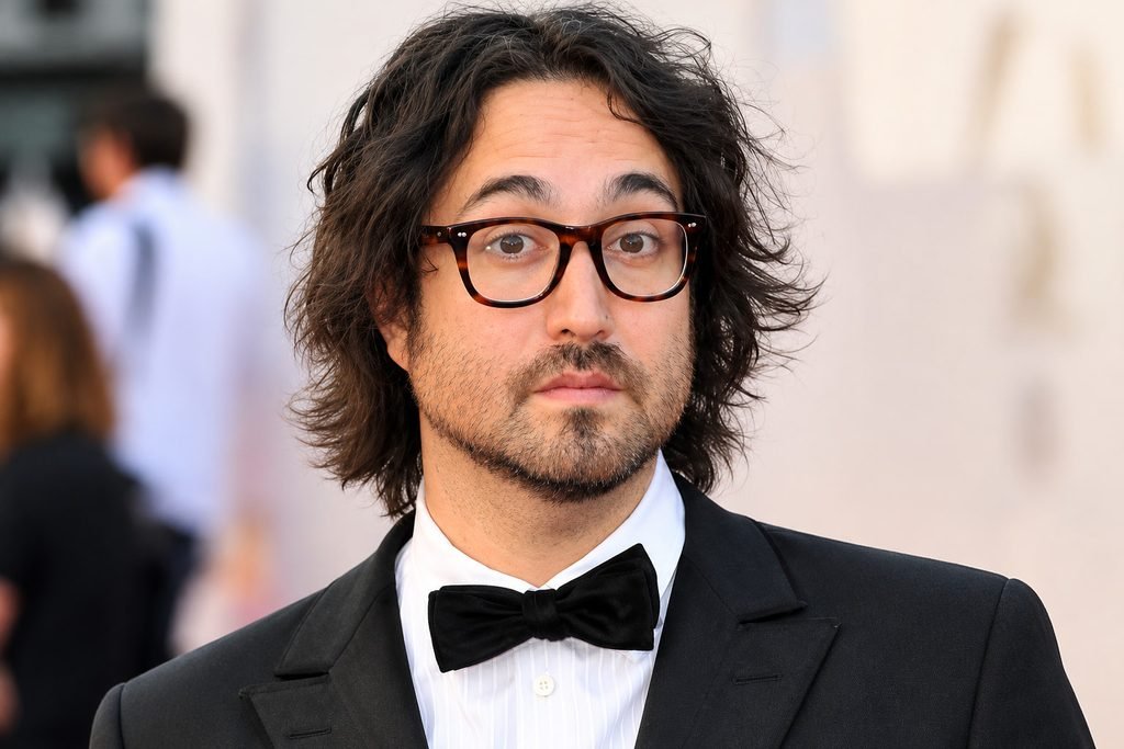 Sean Lennon wearing a black tuxedo, white dress shirt, and black bow tie with glasses at a formal event.