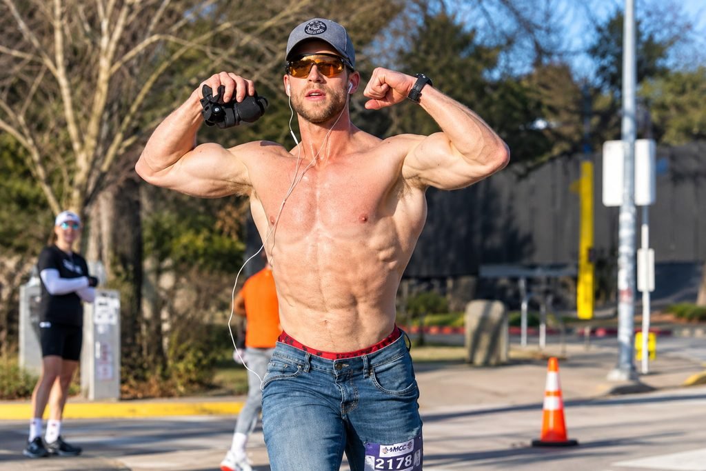 Truett Hanes flexing his muscles outdoors during a race, wearing a cap, sunglasses, and blue jeans.